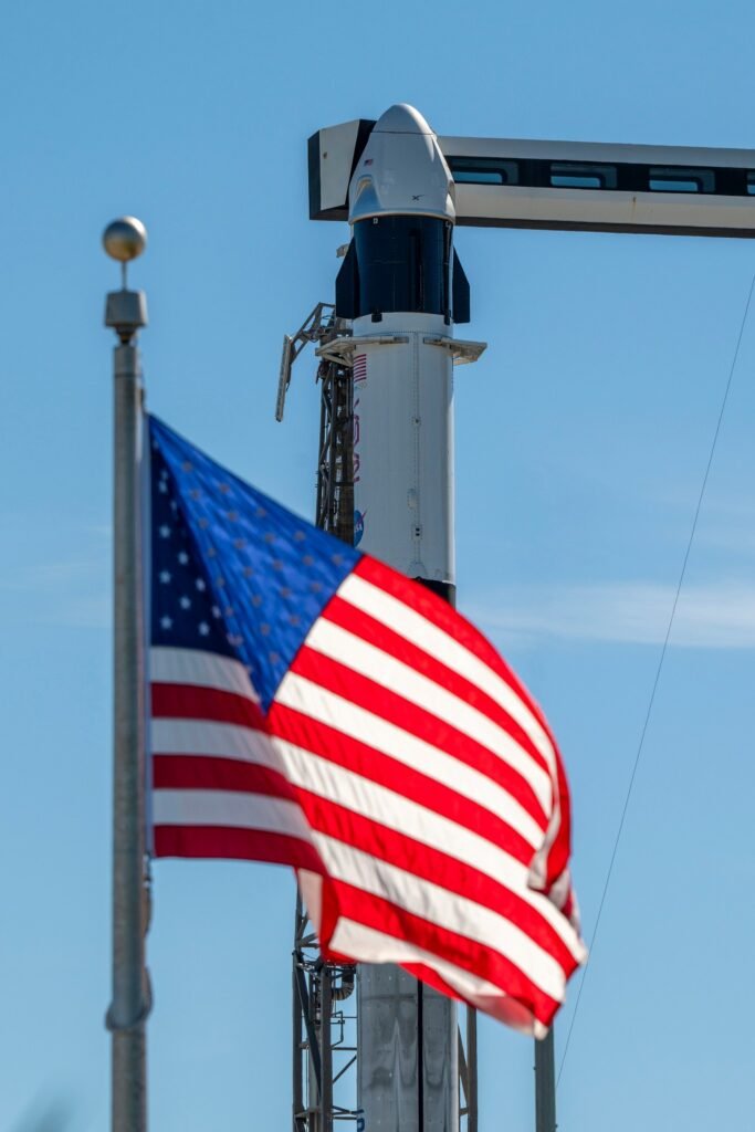 A close-up view of a SpaceX Crew Dragon spacecraft mounted atop a Falcon 9 rocket on the launch pad. The capsule and trunk are connected to ground support hardware, with NASA and mission insignia visible on the rocket’s exterior against a bright blue sky. in the foreground in the American flag.