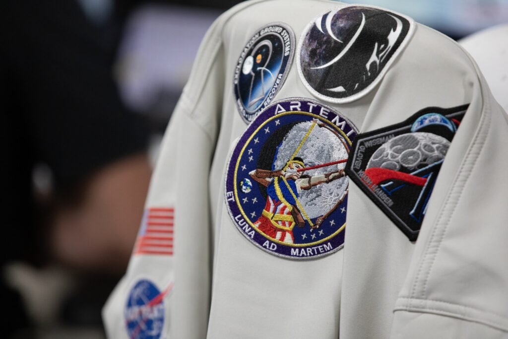 A gray colored jacket with five Artemis mission patches and a NASA insignia hangs on a chair.